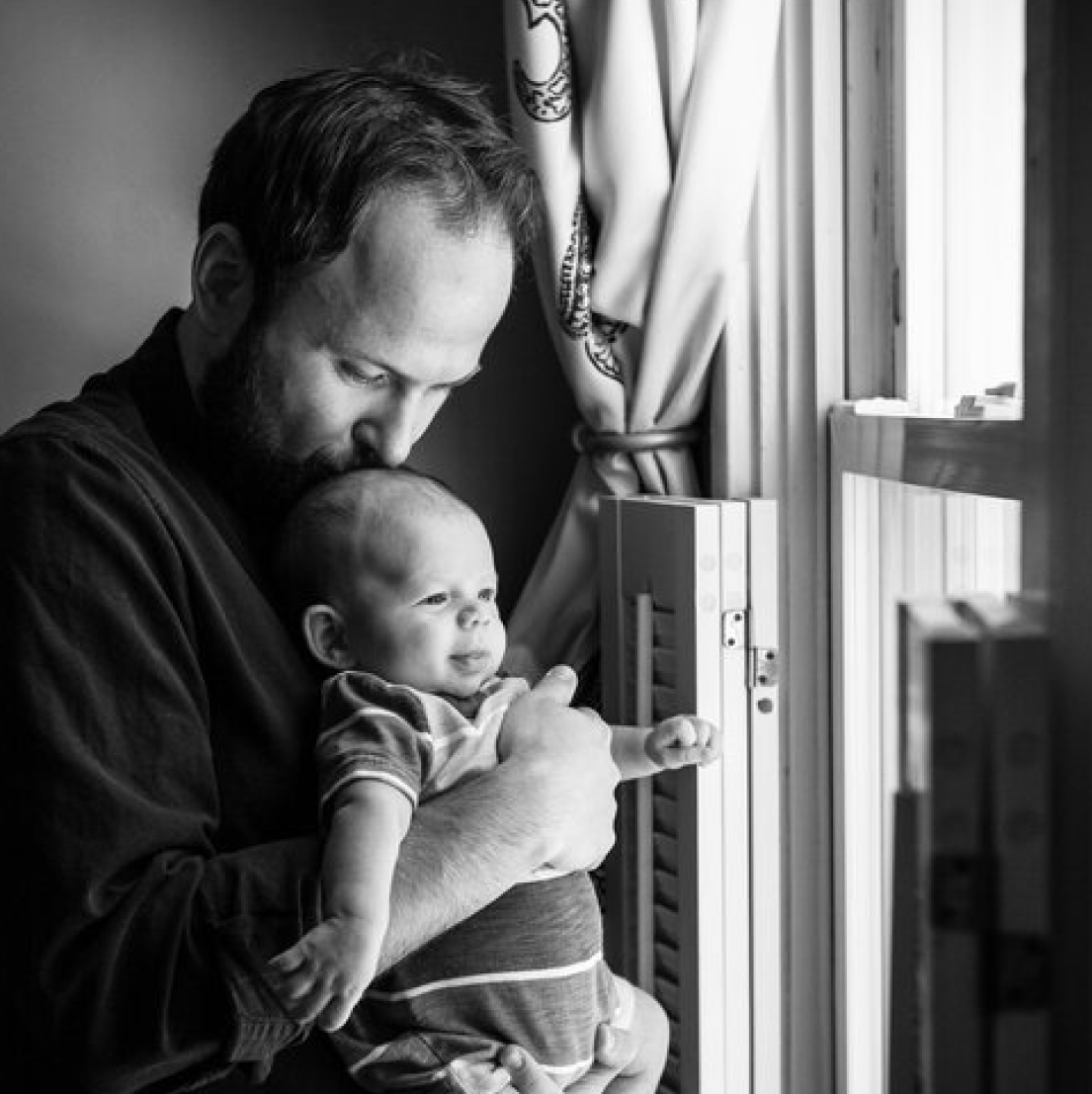 A father hugging and kissing his child as they look out a window in their home.