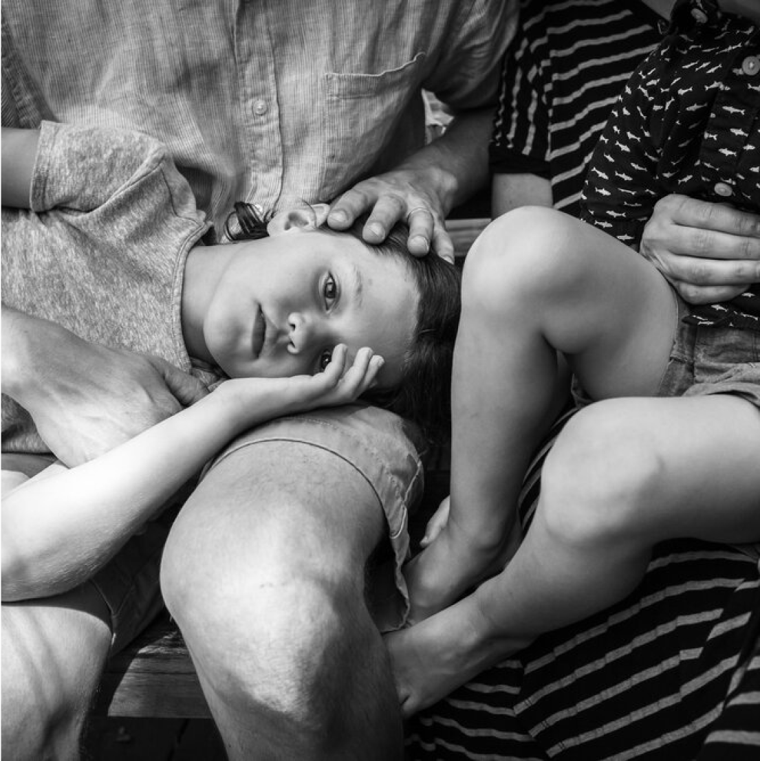 A young boy laying his head on his father's lap with a sibling with their legs curled up sitting next to him.